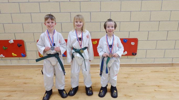 Three children in karate uniforms, each holding a medal and wearing green belts.