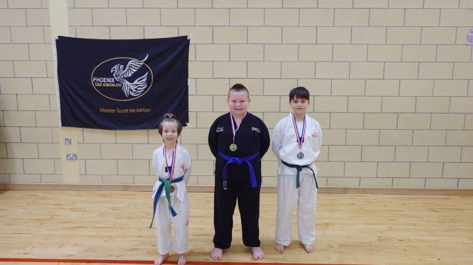 Three young martial artists stand proudly, wearing medals and belts, beside a banner.
